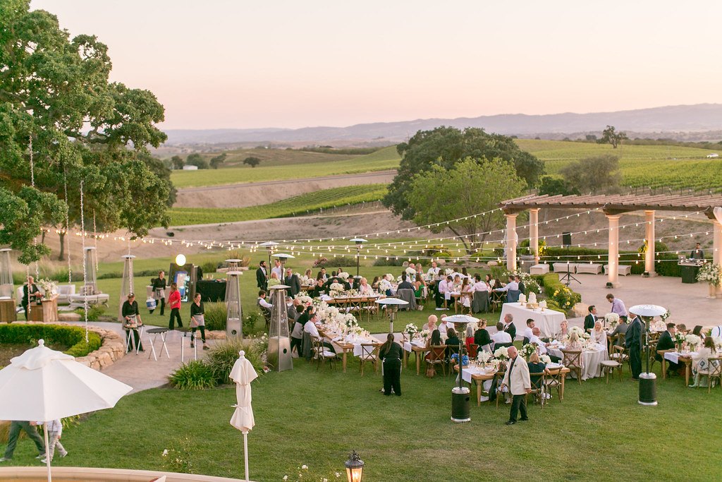 Outdoor evening reception on a lawn under string lights, with guests at long tables and rolling hills beyond